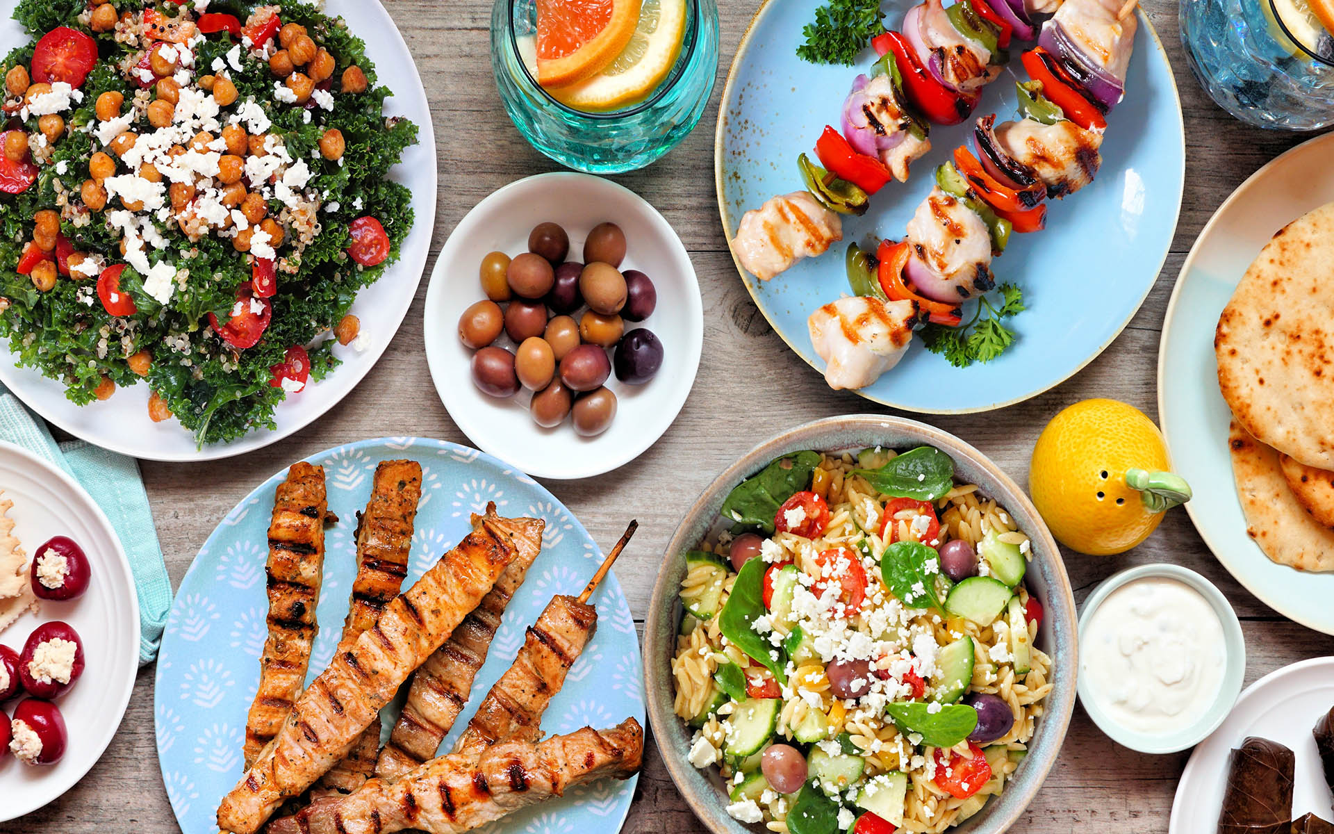 Summer Mediterranean theme food table scene. Top view on a wood banner background. Grilled skewers, salads, snacks, pita and tzatziki.