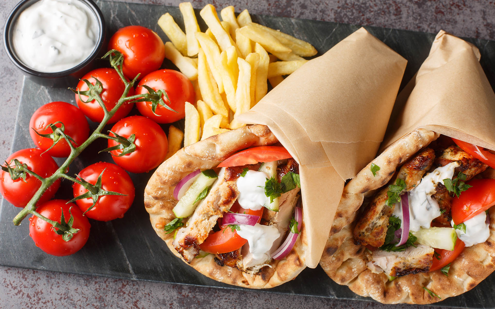 Greek fast food gyros with chicken, tomatoes, cucumbers and onions served with tzatziki sauce and french fries close-up on a board on the table. Horizontal top view from above