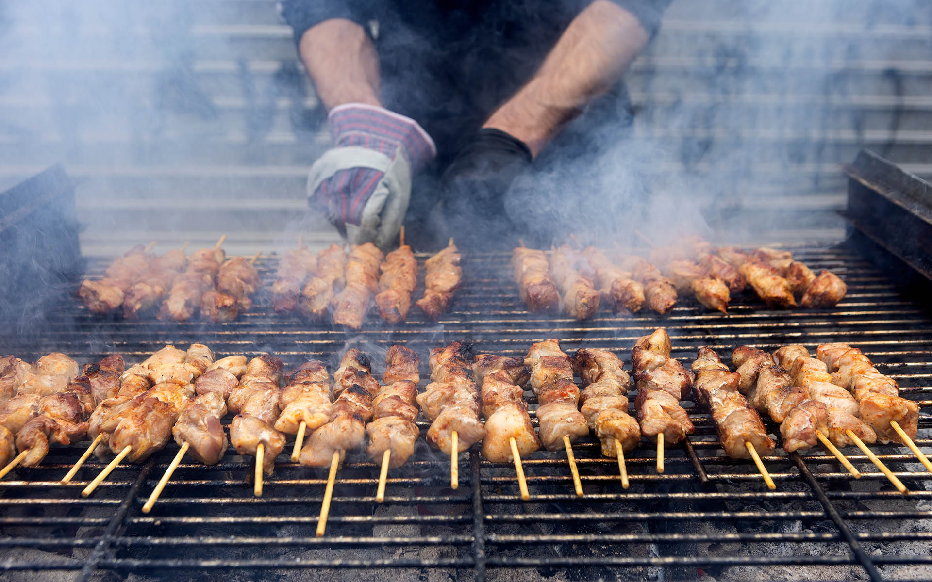 A grill filled with skewers of meat cooking over open flames. The meat browns, while someone tends to the grill wearing gloves. The scene shows a traditional barbecue in progress.Greek Souvlaki
